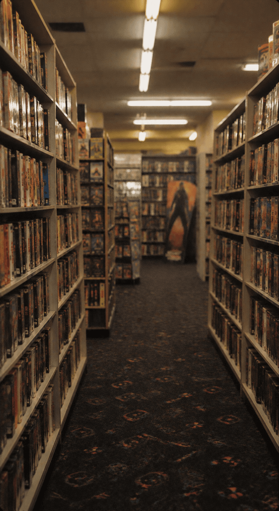 A narrow aisle in a video rental shop, tape-filled shelves on both sides and a figure on a poster at the far end.